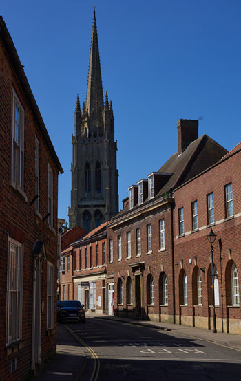 The image shows a street scene in Louth, Lincolnshire, with the prominent spire of St James' Church rising above the surrounding buildings. The photograph captures the urban architecture typical of this historic market town, displaying red-brick facades and arched windows under a clear blue sky. Taken in late morning during spring, the lighting highlights the intricate details of the church's Gothic architecture as well as the texture of the brickwork along the street. St James' Church serves as the main subject and recognizable landmark in this urban landscape, illustrating the cultural and architectural character of Louth in Lincolnshire during the spring season.